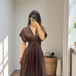 May include: A brown midi dress with a V-neckline and short sleeves. The dress features a tie waist and a flowing skirt. The person in the image is wearing sandals. The dress is made of a soft, flowing fabric.