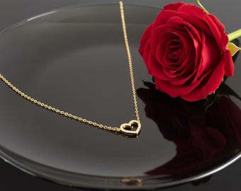 Può includere: Una collana dorata con un ciondolo a forma di cuore, posta su un piatto nero accanto a una rosa rossa. La collana ha una catena sottile e il ciondolo a forma di cuore è adornato con piccole pietre scintillanti. La rosa è in piena fioritura.