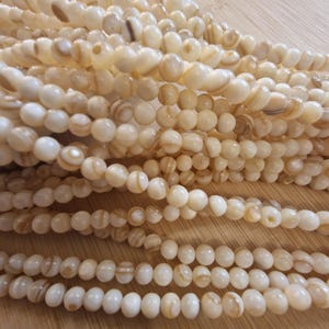 Peut inclure: Une collection de perles rondes blanc cassé avec des motifs naturels tourbillonnants de couleur beige et marron. Les perles sont disposées en plusieurs rangées parallèles, présentant une finition lisse et polie ainsi qu'une forme sphérique.