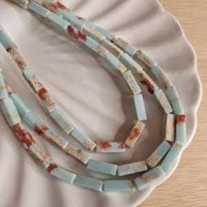 Pode incluir: Um colar de várias camadas com contas retangulares em azul-pálido com inclusões naturais em tons de castanho-avermelhado e creme. As contas estão dispostas numa série de laços em cascata, apresentando um acabamento liso e polido em todo o design.