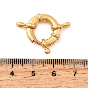 Puede incluir: Un cierre de resorte circular de tono dorado con dos anillas laterales y una pequeña lengüeta esférica. El cierre está colocado sobre una regla de madera, mostrando una anchura de aproximadamente 1,9 cm de anilla a anilla para este componente.