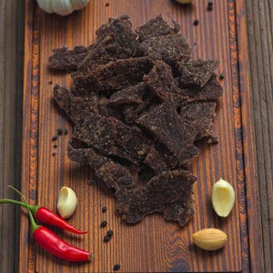 May include: A wooden cutting board displays a pile of dark brown beef jerky. Red chili peppers, garlic cloves, peppercorns, and a white bulb of garlic are arranged around the jerky, creating a rustic food presentation.