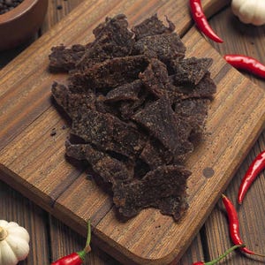 May include: Close-up of a wooden cutting board with a pile of dark brown beef jerky. Red chili peppers, garlic, and peppercorns are scattered around the board on a wooden surface. The jerky appears to be seasoned.