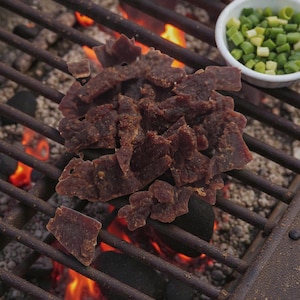 May include: A pile of dark brown beef jerky sits on a metal grill over a bed of hot coals and flames. Two small white bowls hold red sauce and chopped green onions. The image captures a grilling scene.