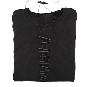K&ouml;nnte beinhalten: Ein schwarzes Langarmshirt mit einer vertikalen Reihe aus zehn dunklen, texturierten, spindelf&ouml;rmigen Perlen, die mittig auf der Brust durch d&uuml;nne schwarze Kordeln befestigt sind, welche zum Halsausschnitt f&uuml;hren und in einem kleinen Verschluss enden.