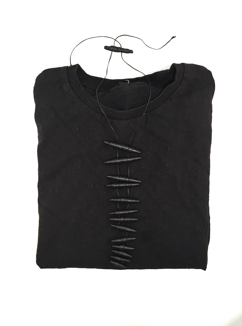 K&ouml;nnte beinhalten: Ein schwarzes Langarmshirt mit einer vertikalen Reihe aus zehn dunklen, texturierten, spindelf&ouml;rmigen Perlen, die mittig auf der Brust durch d&uuml;nne schwarze Kordeln befestigt sind, welche zum Halsausschnitt f&uuml;hren und in einem kleinen Verschluss enden.