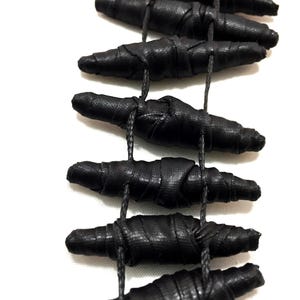 K&ouml;nnte beinhalten: Eine Halskette mit mehreren l&auml;nglichen, zylindrischen schwarzen Perlen, die mit einem strukturierten Stoff umwickelt sind. Die Perlen sind in einer vertikalen, leiterartigen Formation angeordnet und durch d&uuml;nne schwarze Kordeln miteinander verbunden.