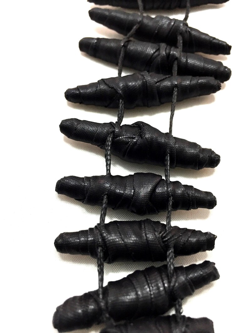 K&ouml;nnte beinhalten: Eine Halskette mit mehreren l&auml;nglichen, zylindrischen schwarzen Perlen, die mit einem strukturierten Stoff umwickelt sind. Die Perlen sind in einer vertikalen, leiterartigen Formation angeordnet und durch d&uuml;nne schwarze Kordeln miteinander verbunden.