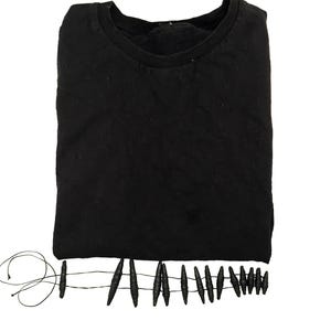 K&ouml;nnte beinhalten: Ein einfarbiges schwarzes T-Shirt mit Rundhalsausschnitt. Am unteren Saum befindet sich ein dekoratives Fransenelement, das aus einer d&uuml;nnen schwarzen Schnur besteht, die durch eine Reihe kleiner, l&auml;nglicher, strukturierter schwarzer Perlen verl&auml;uft.