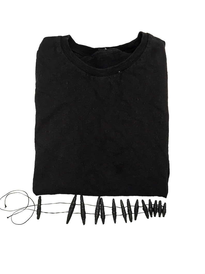 K&ouml;nnte beinhalten: Ein einfarbiges schwarzes T-Shirt mit Rundhalsausschnitt. Am unteren Saum befindet sich ein dekoratives Fransenelement, das aus einer d&uuml;nnen schwarzen Schnur besteht, die durch eine Reihe kleiner, l&auml;nglicher, strukturierter schwarzer Perlen verl&auml;uft.
