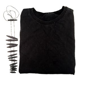 K&ouml;nnte beinhalten: Ein einfarbig schwarzes Langarmshirt mit Rundhalsausschnitt, kombiniert mit einer langen Halskette aus einer d&uuml;nnen schwarzen Schnur und mehreren kleinen, dunklen, l&auml;nglichen, spindelf&ouml;rmigen Perlen, die in einer Reihe angeordnet sind.