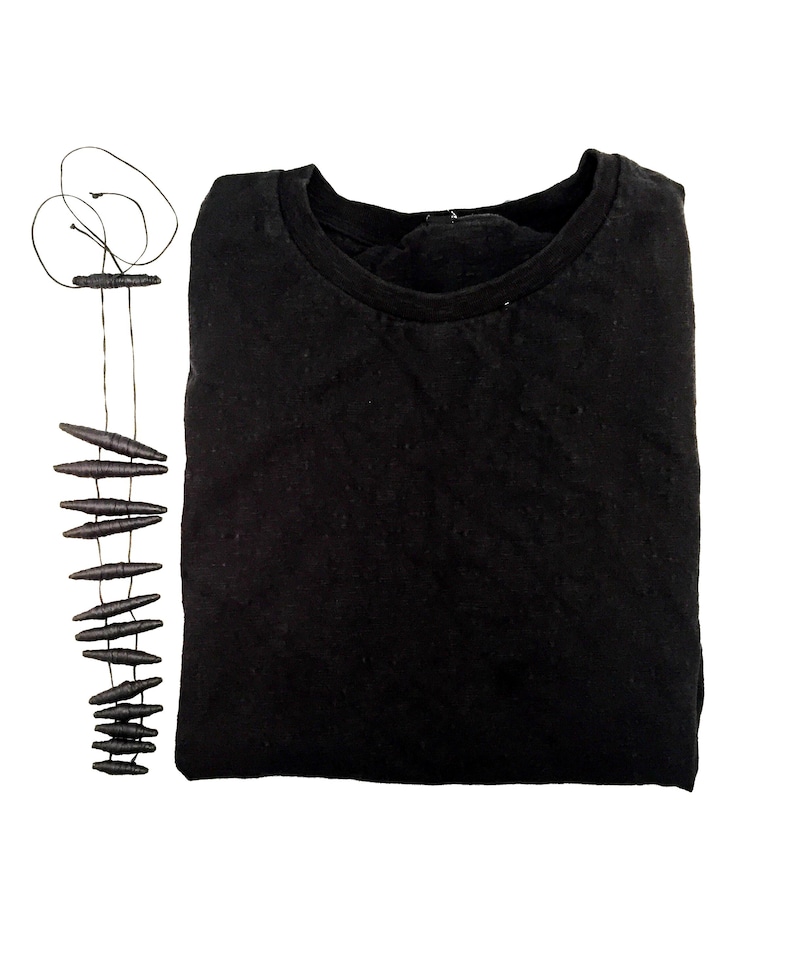 K&ouml;nnte beinhalten: Ein einfarbig schwarzes Langarmshirt mit Rundhalsausschnitt, kombiniert mit einer langen Halskette aus einer d&uuml;nnen schwarzen Schnur und mehreren kleinen, dunklen, l&auml;nglichen, spindelf&ouml;rmigen Perlen, die in einer Reihe angeordnet sind.