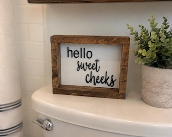 May include: A rectangular wooden sign with a dark brown stained frame and a white background. The sign features black, raised, three-dimensional lettering that reads "hello sweet cheeks" in a mix of sans-serif and cursive script fonts.