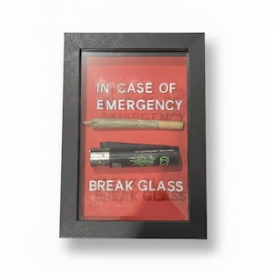 May include: A framed display with a red background, featuring a pre-rolled joint and a black lighter. The text "IN CASE OF EMERGENCY" and "BREAK GLASS" are printed on the glass.
