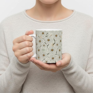 Pode incluir: Caneca de cerâmica branca com um padrão de margaridas com centros amarelos e folhas cinzentas sobre um fundo bege claro. A caneca tem uma alça branca e é segurada por uma pessoa que usa um suéter cinza claro.