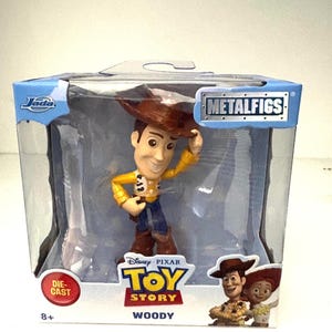 May include: A die-cast metal Woody figure from the Disney Pixar Toy Story movie, packaged in a clear plastic box. The figure is wearing a cowboy hat, yellow shirt, blue pants, and brown boots. The box has the Metalfigs logo and the Jada logo.