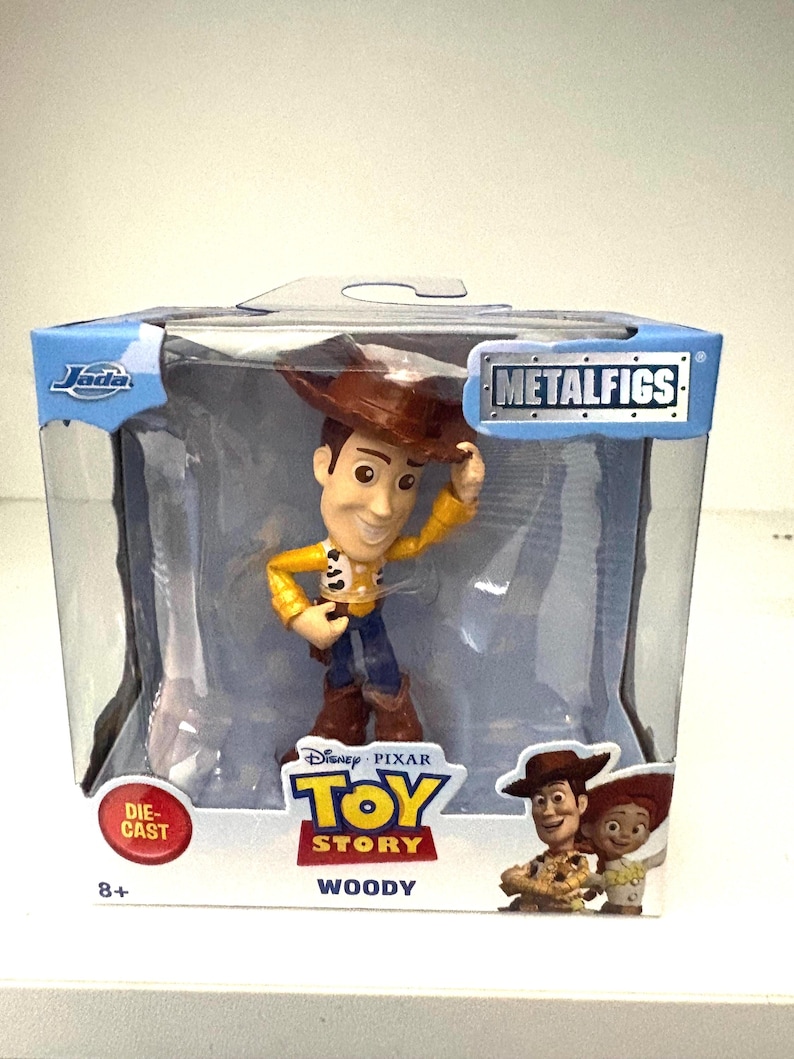 May include: A die-cast metal Woody figure from the Disney Pixar Toy Story movie, packaged in a clear plastic box. The figure is wearing a cowboy hat, yellow shirt, blue pants, and brown boots. The box has the Metalfigs logo and the Jada logo.