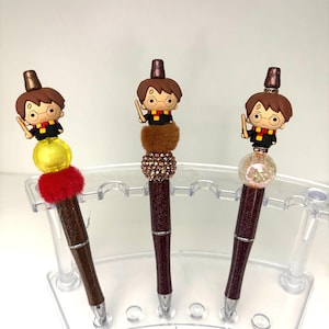 May include: Three decorative pens featuring a cartoon character with glasses, a wand, and a brown hat. The pens have brown glitter barrels and are displayed in a clear plastic holder. Each pen has a different decorative element: yellow, brown, and pink.
