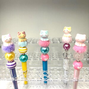 May include: A collection of decorative pens featuring cat-shaped toppers. Each pen has a unique design with colorful beads, pom-poms, and a cat figurine. The pens are displayed in a clear plastic organizer.