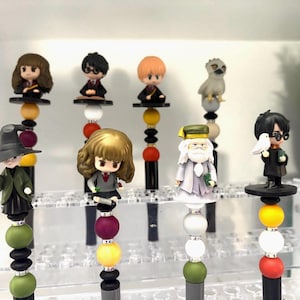 May include: A collection of decorative pens featuring miniature figurines of characters from a fantasy series. Each pen has a unique design with colorful beads and a character figure on top. The pens are displayed in a clear acrylic holder.