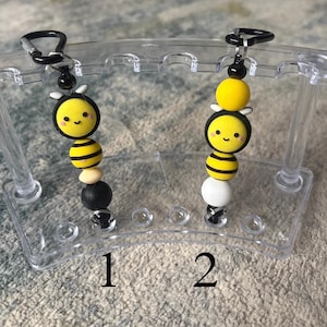 May include: Two beaded bee keychains. Each keychain features a smiling bee charm with yellow and black stripes, a black clasp, and various colored beads. The keychains are displayed on a clear plastic stand.