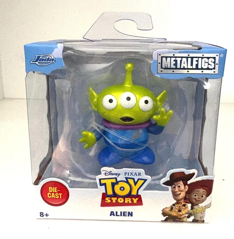 May include: A die-cast Toy Story Alien figure in a clear plastic package. The green alien has three eyes, a purple collar, and blue clothing. The package includes the Toy Story logo, the word "Alien", and images of Woody and Jessie.