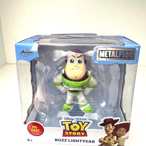 May include: A die-cast Buzz Lightyear figure from the Disney Pixar Toy Story franchise. The figure is in a clear plastic display case with the words "METALFIGS" and "Buzz Lightyear" printed on the packaging. The figure is wearing a green and white spacesuit.