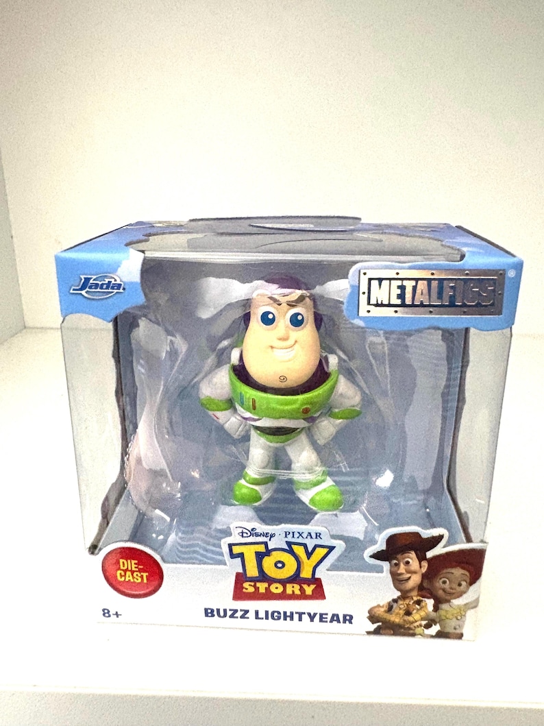 May include: A die-cast Buzz Lightyear figure from the Disney Pixar Toy Story franchise. The figure is in a clear plastic display case with the words "METALFIGS" and "Buzz Lightyear" printed on the packaging. The figure is wearing a green and white spacesuit.