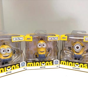 Puede incluir: Tres figuras de Minions de metal fundido Jada Toys Metalfigs en cajas con ventana amarillas y blancas. Las figuras son Otto, Kevin y Stuart, cada una con peto azul. El embalaje presenta el logotipo de Minions, la marca Metalfigs y el nombre del personaje impreso en la parte frontal de cada caja.