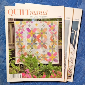 May include: Three issues of "QUILTmania" magazine are stacked on a blue surface. The cover of the main magazine features a colorful quilt with a floral design. The magazine's title is in a coral color. The other two magazines have issue numbers 135 and 138.