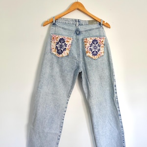 Op de afbeelding: Lichtblauwe jeans met bloemen geborduurde achterzakken. De jeans hangen aan een houten hanger tegen een witte achtergrond. De jeans hebben een rechte pijp en een rafelige zoom.