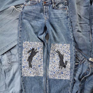 Könnte beinhalten: Hellblaue Jeans mit Aufnähern mit floralem Muster und schwarzen Tiersilhouetten. Die Jeans haben ein Sterndesign auf der Vordertasche und sind von anderen Jeanskleidungsstücken umgeben.