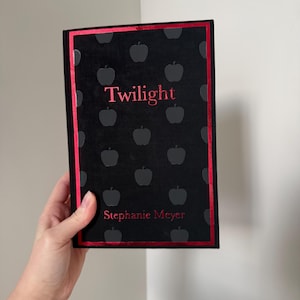 May include: A black book with the title "Twilight" in red lettering. The cover features a pattern of grey apple silhouettes and a red border. The author's name, Stephanie Meyer, is also in red.