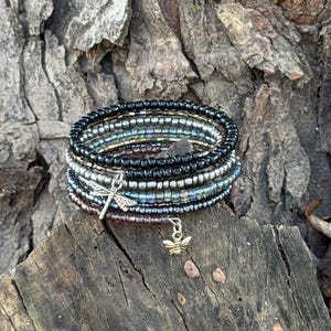May include: A multi-strand wrap bracelet composed of small glass seed beads in shades of black, metallic silver, and deep purple. The bracelet features two dangling metal charms, one shaped like a dragonfly and the other shaped like a bee.