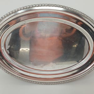 Peut inclure: Plateau de service ovale argenté avec une bordure en forme de corde. Le plateau a une surface réfléchissante et les bords sont légèrement surélevés. Le design est simple et élégant, adapté au service ou à l'exposition.