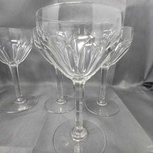 Può includere: Un gruppo di calici da vino in cristallo trasparente. I calici presentano un design classico con una coppa rotonda, un lungo stelo e una base circolare. Le coppe sono decorate con un motivo inciso. I calici sono disposti su una superficie grigia.