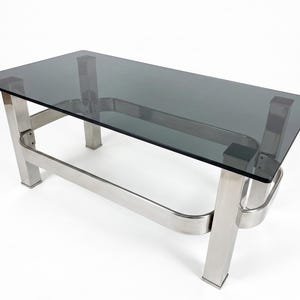 Könnte beinhalten: Ein rechteckiger Couchtisch mit einer dunkelgrau getönten Glasplatte, die von einem Rahmen aus gebürstetem silbernem Metall getragen wird. Das Untergestell besteht aus vier vertikalen Beinen, die durch eine abgerundete Metallstrebe verbunden sind.
