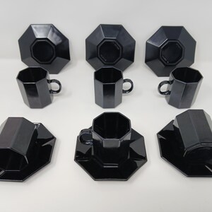 Peut inclure: Un ensemble de six tasses à thé et soucoupes noires de forme géométrique. Les tasses ont une anse et reposent sur des soucoupes octogonales assorties. La finition brillante reflète la lumière. L'ensemble est sur une surface blanche.