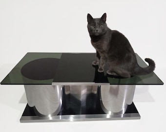 Op de afbeelding: Een rechthoekige salontafel met een donker glazen blad, een cirkelvormige zwarte inleg en een massief zwart middenpaneel. Het onderstel bestaat uit drie gepolijste zilverkleurige metalen cilinders op een zwart en zilverkleurig rechthoekig platform.