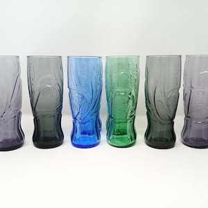 Può includere: Sei bicchieri alti e colorati. I bicchieri sono nelle tonalità del viola, grigio, blu e verde. Ogni bicchiere ha un design unico ed è fatto di vetro. I bicchieri sono disposti in fila su uno sfondo bianco.