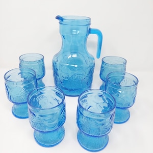 Può includere: Un set di cristalleria blu traslucido composto da una brocca con manico e sei bicchieri abbinati. Ogni pezzo presenta un motivo in rilievo stampato di frutta e foglie sulla superficie esterna, con un colore blu brillante e uniforme.