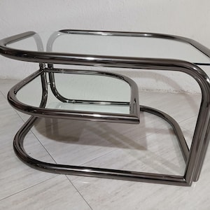 Op de afbeelding: Een moderne salontafel met twee niveaus, met een helder glazen blad en een onderste plank. Het frame is gemaakt van gebogen, donker metaal. De tafel heeft een uniek, geometrisch ontwerp, geschikt voor een woonkamer of lounge.