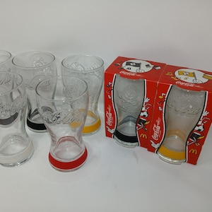 Può includere: Una collezione di bicchieri Coca-Cola trasparenti con basi colorate, tra cui rosso, giallo e nero. I bicchieri sono esposti con una scatola rossa con marchio Coca-Cola e illustrazioni di cartoni animati. I bicchieri hanno un design classico Coca-Cola.