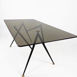 Puede incluir: Una mesa de comedor rectangular con un tablero de cristal ahumado y esquinas redondeadas. La mesa se apoya en cuatro patas finas de metal negro inclinadas hacia fuera, cada una con un pequeño pie esférico de color latón en la base.