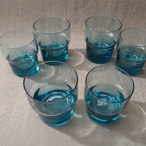 Op de afbeelding: Een set van zes bijpassende drinkglazen gemaakt van doorschijnend blauwgroen glas. Elk glas heeft een eenvoudige cilindrische vorm met een iets bredere rand en een gladde, heldere bodem. De glazen zijn gerangschikt in drie rijen.