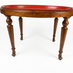 Peut inclure: Une table ronde en bois avec un plateau rouge vif et lisse. La table possède quatre pieds tournés avec une finition brun foncé, chacun se terminant par une petite roulette en métal arrondie. Le design inclut un rebord surélevé.