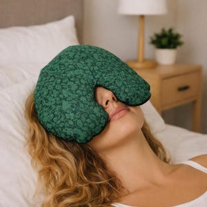 May include: A green eye pillow with a floral pattern rests on a person's face. The pillow is dark green with a black border. The person is lying on a white bed with a white pillow and a wooden nightstand in the background.