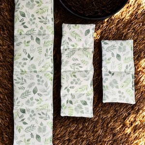Peut inclure: Trois coussins chauffants en tissu rectangulaires de longueurs variables, pr&eacute;sentant un fond blanc avec un motif r&eacute;p&eacute;titif de feuilles et de feuillage verts. Les coussins sont segment&eacute;s en sections par des coutures horizontales.