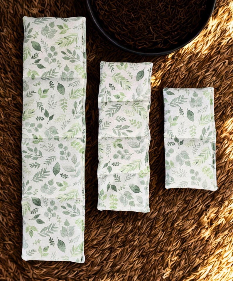 Peut inclure: Trois coussins chauffants en tissu rectangulaires de longueurs variables, pr&eacute;sentant un fond blanc avec un motif r&eacute;p&eacute;titif de feuilles et de feuillage verts. Les coussins sont segment&eacute;s en sections par des coutures horizontales.