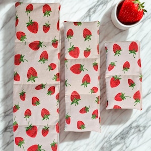 May include: Three fabric items with a strawberry pattern. The strawberries are red with green stems, scattered on a light pink background. A small white bowl holds a fresh strawberry. The items appear to be reusable food wraps.